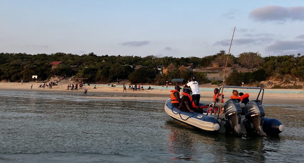 A motorboat with six people aboard approaches a beach full of people, with buildings visible among the trees