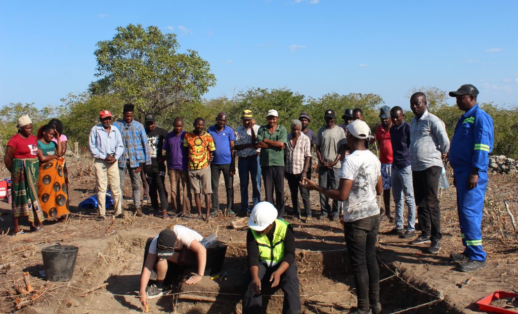 A group of people stand around an archaeological trench with two archaeologists excavating and a third archaeologist speaking to the group.